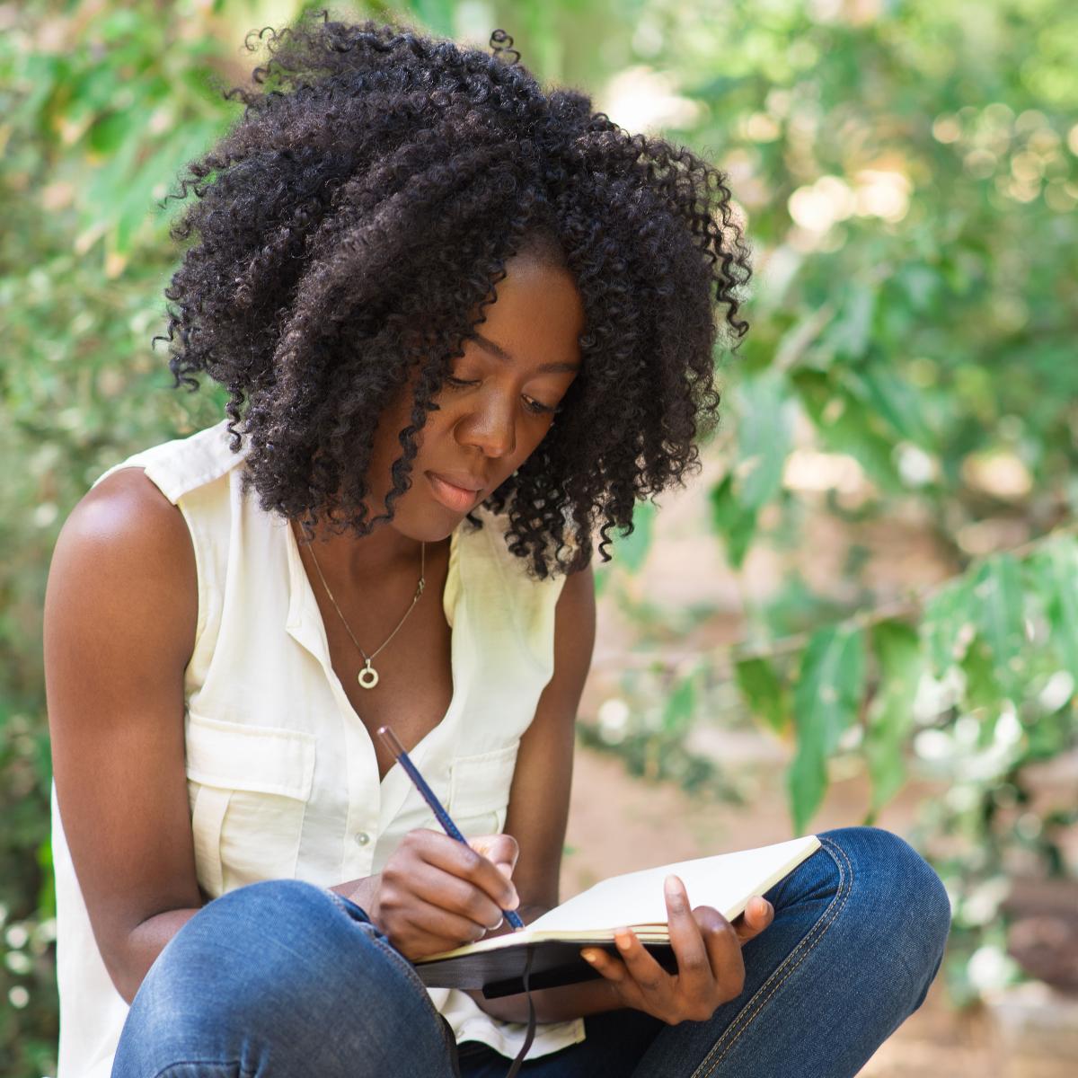 Young adult sitting cross-legged outside while writing in a journal