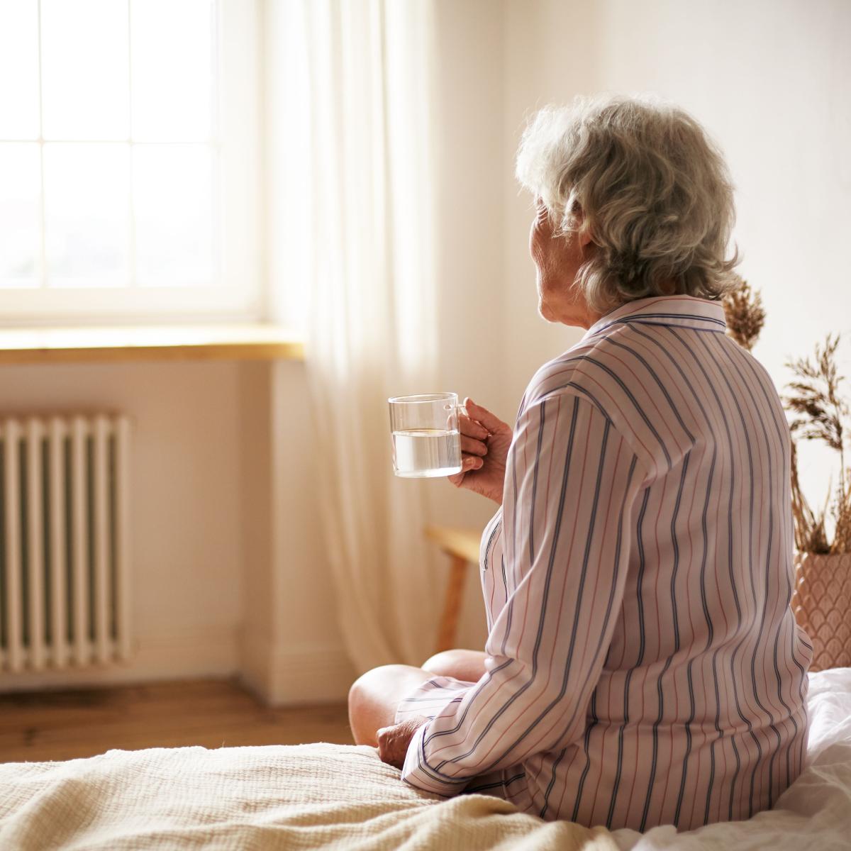 Older Adult in Pajamas Sitting on Bed, Looking Out a Window, While Drinking a Hot Beverage