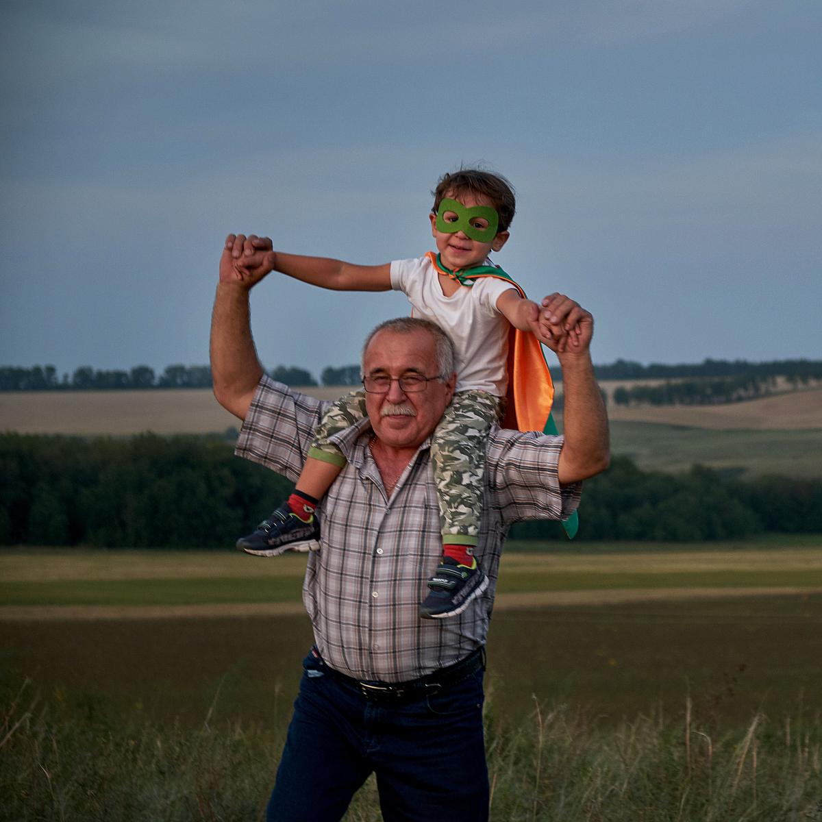 Grandparent holding young grandson, who is wearing a mask and cape, on shoulders 